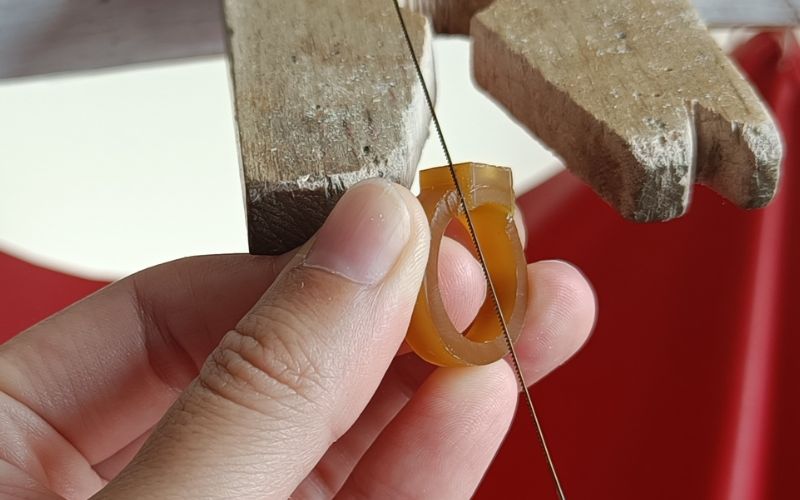 Close up of hands holding a piece of gold ring wax. Piercing up top next to a setting part and because the saw frame is angled it's not piercing the bottom part