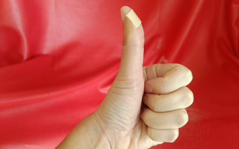 Hand doing thumbs up in front of a red background with a bandaid on the thumb