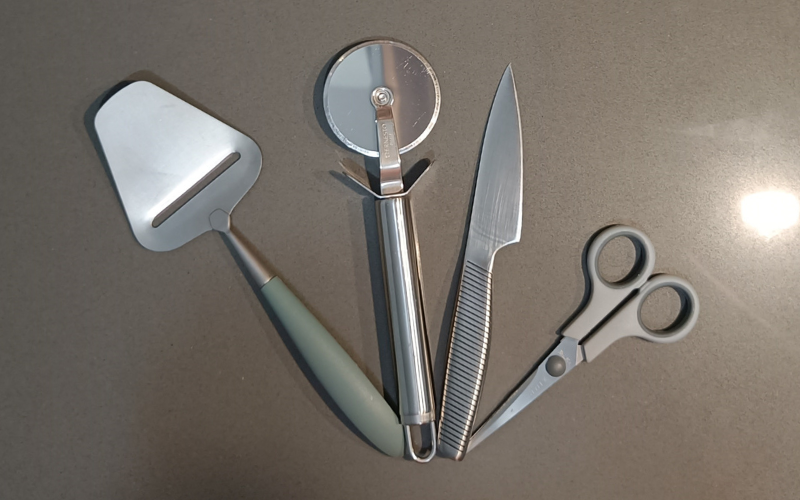 cheese slicer, pizza cutter, sharp small knife and scissors fanned out on a grey surface 