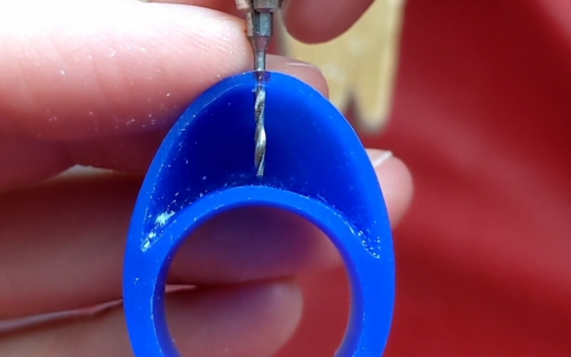 Close up of hands holding blue wax wing with an arch shaped opening on top of the ring, a drillbit goes through the center of the opening