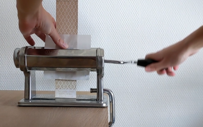 Pasta roller, left hand os feeding texture paper with sheet wax through the rollers, right hand is using the handle to roll it through