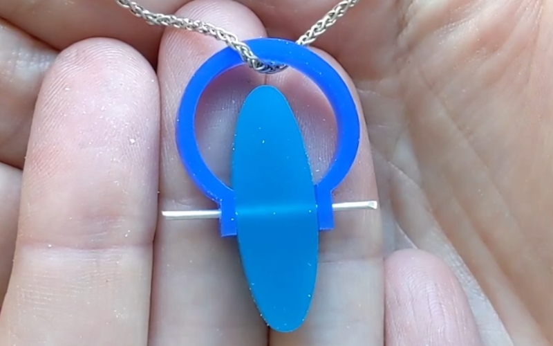 Close up of hands holding a blue wax ring with a central oval section from turquoise wax. The center section is swiveled so it sits flat and a chain goes through the ring band. 