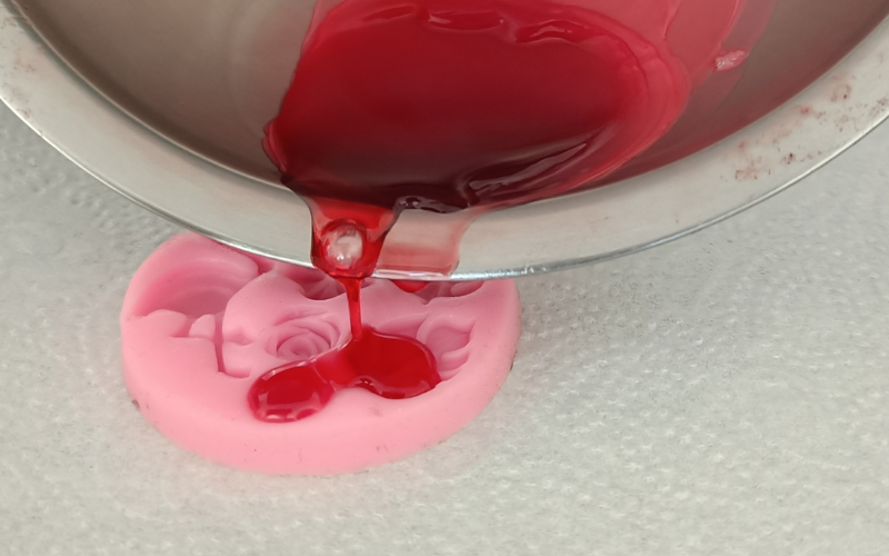 Close up of a pink silicone mold with red wax being poured inside from a pan