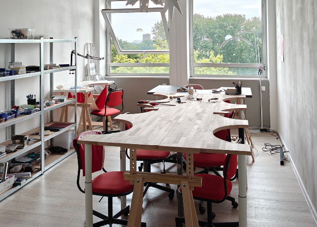 Studio space with big windows in the back, Big table with 6 jewellery benches is in the middle. 2 smaller jewellery benches are at the end and to the left side. 2 big cabinets with storage are next to the left bench.