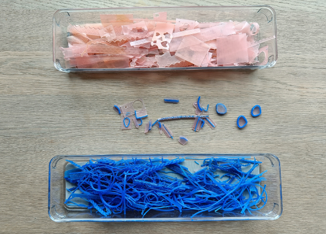 Table with 2 plastic trays, one filled with blue wire wax and one with pink sheet wax. In the middle scrap pieces of wax that have both and need to be sorted still