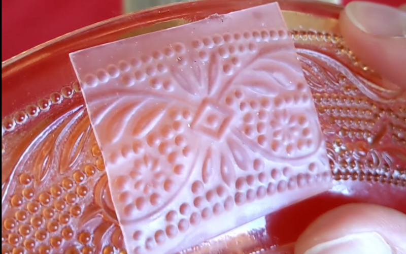 Close up of glass plate with pink sheet wax pushed against the pattern, you can see that the pattern has transferred to the sheet wax