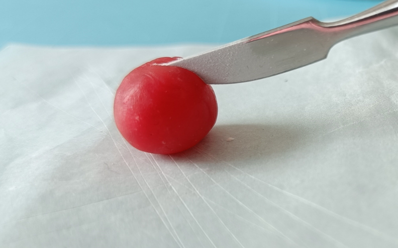 Close up of a ball of red wax on top of paper. A knife carving tool is slicing in the wax. The blade of the carving tool is covered in talcum powder.  