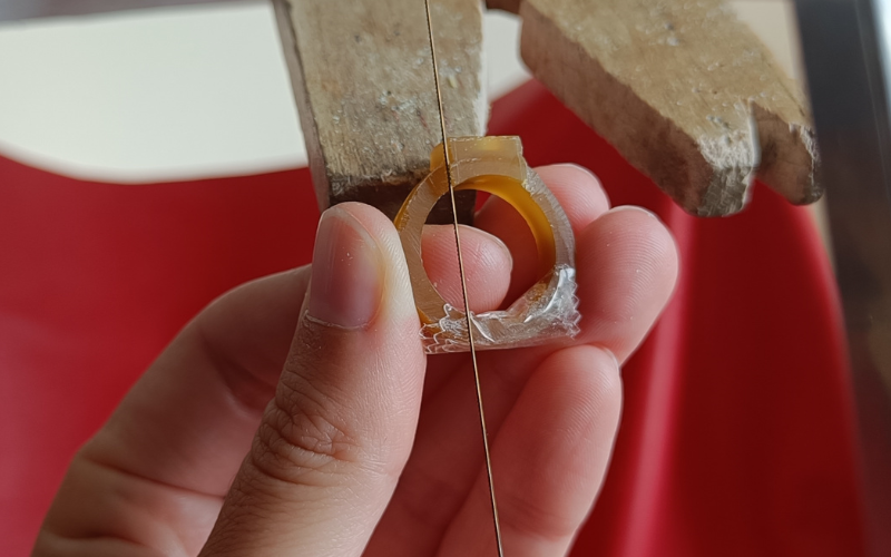 Close up of hands holding a piece of gold ring wax. Piercing up top next to a setting part there's tape on the bottom part to remind you to angle the saw frame