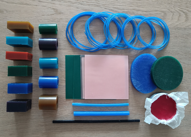 Flat lay of all the wax that's included. Blocks of wax in blue, orange, green, turquoise, purple and gold. Ring wax in blue, gold, green, turquoise and purple. Green and pink sheet wax, 5 coils of wax wire. Blue oval bangle wax, green round bangle wax. Red wax in a tin. 2 blue wax tubes, dark blue square wire