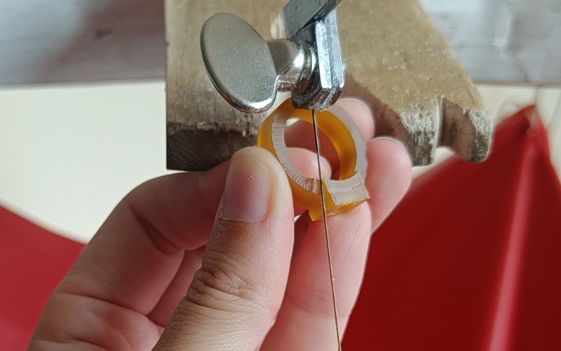 Close up of hands holding a piece of gold ring wax. The ring is held upside down so teh majority is facing away from the saw frame. 